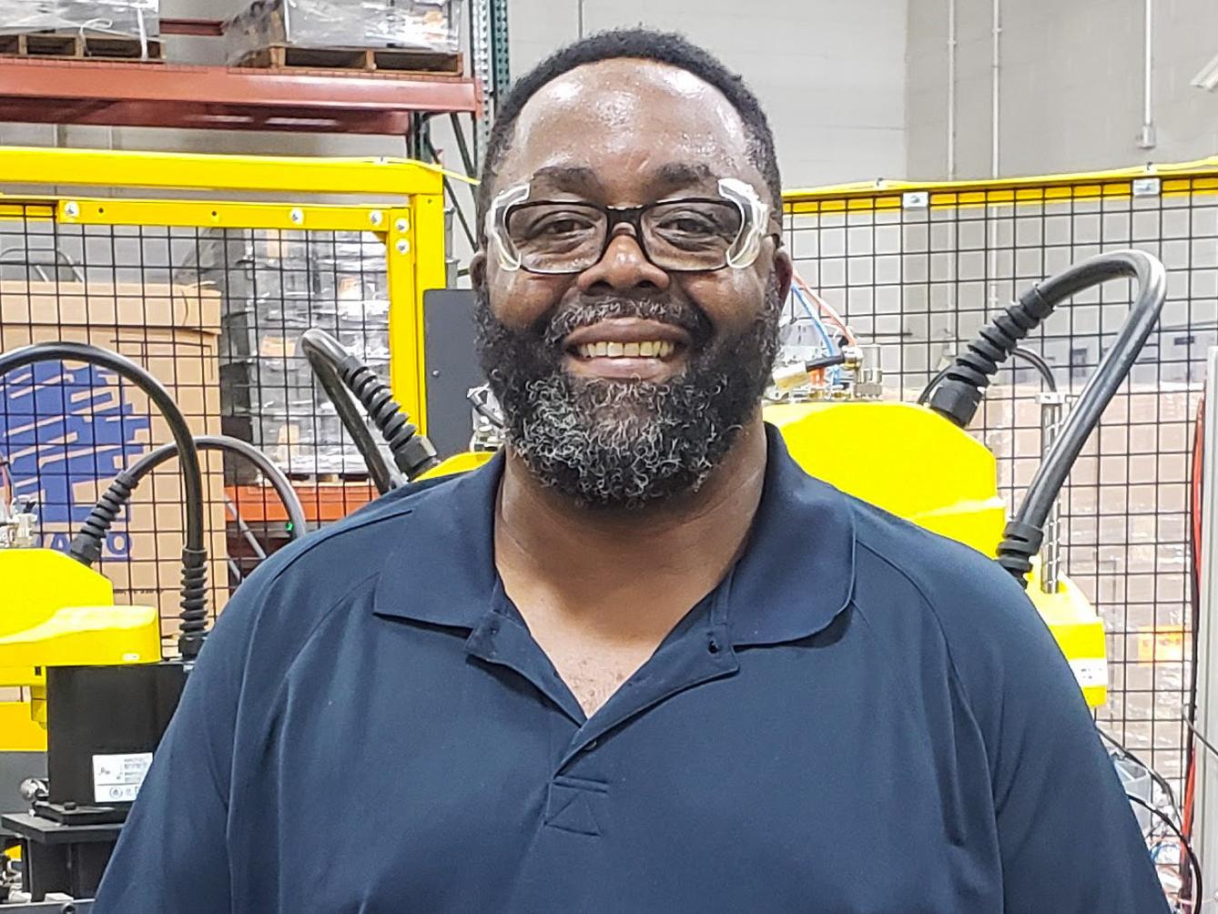 Ryan Burton in a navy blue shirt stands in front of a yellow piece of equipment in a factory.