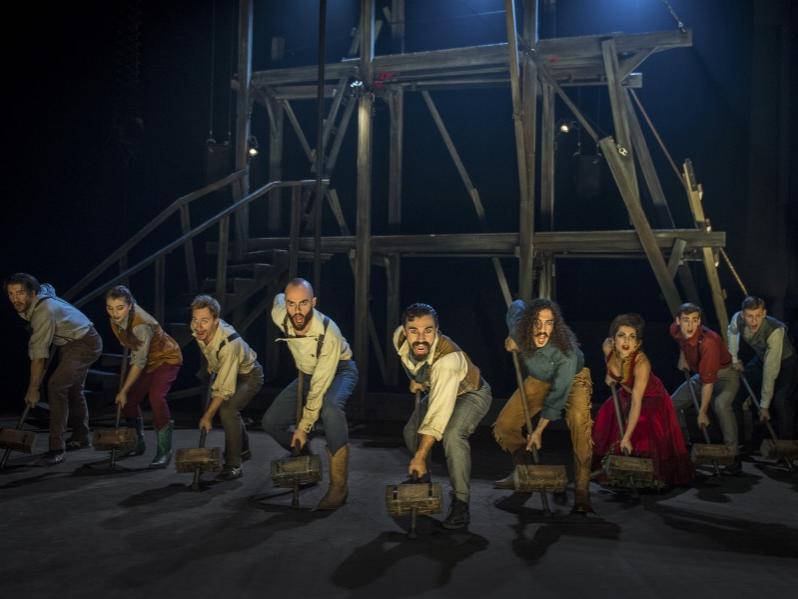Nine acrobats dressed in 1800s-era clothing stand in ​“V” formation in front of wood scaffolding while wielding wooden-block hammers.