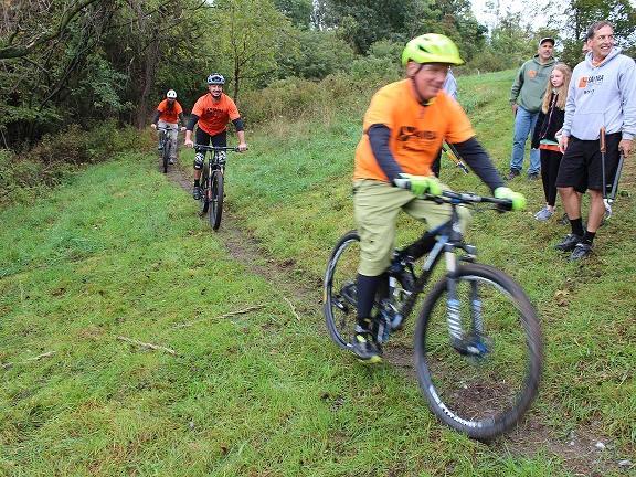 Three men wearing bike helmets and T-shirts ride mountain bikes on a narrow, dirt trail. Four people watch them on either side of the trail. One man is holding garden shears.
