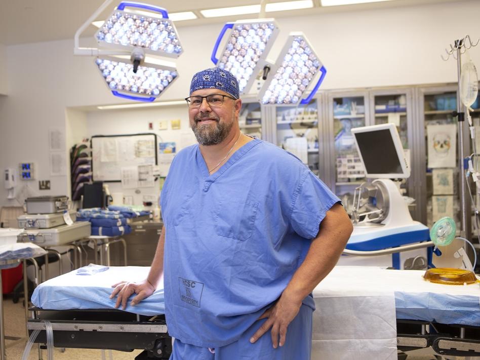 Glenn Weidler leans against an operating room table. He is wearing scrubs and a pair of glasses.