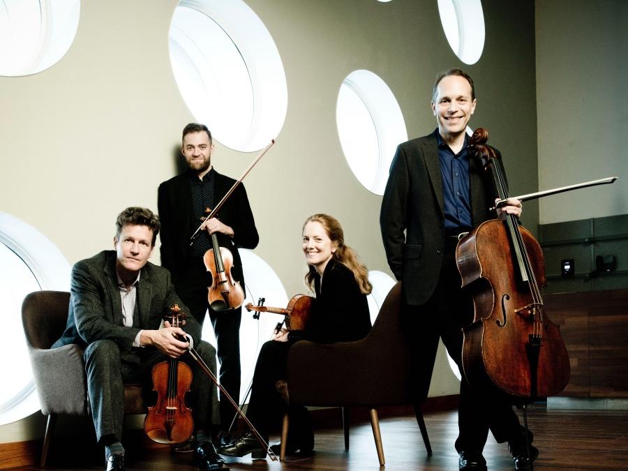 Four musicians sit and lean on modern furniture while they hold their instruments.