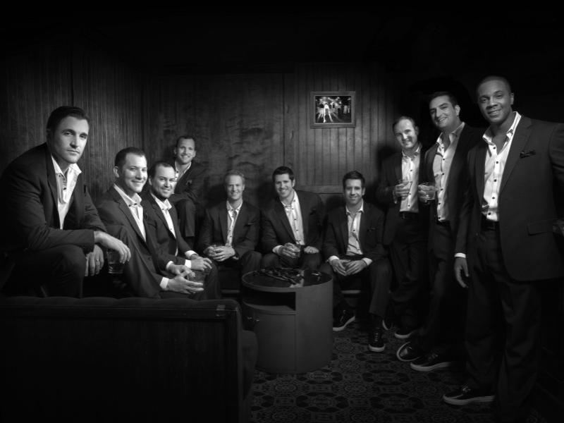 Each of the 10 singers of a cappella group Straight No Chaser wear a suit and sneakers and hold a drink while they smile at the camera.