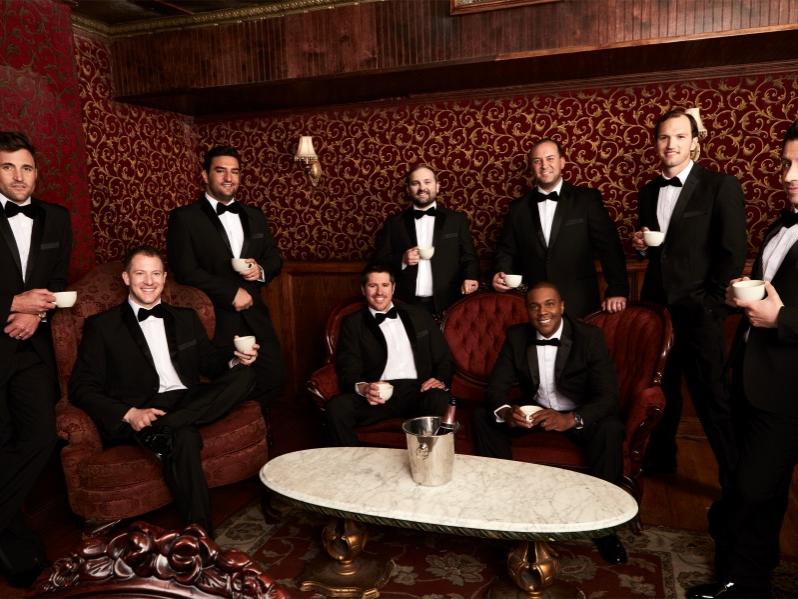 Nine formally dressed men, some sitting and some standing, smile for the camera.