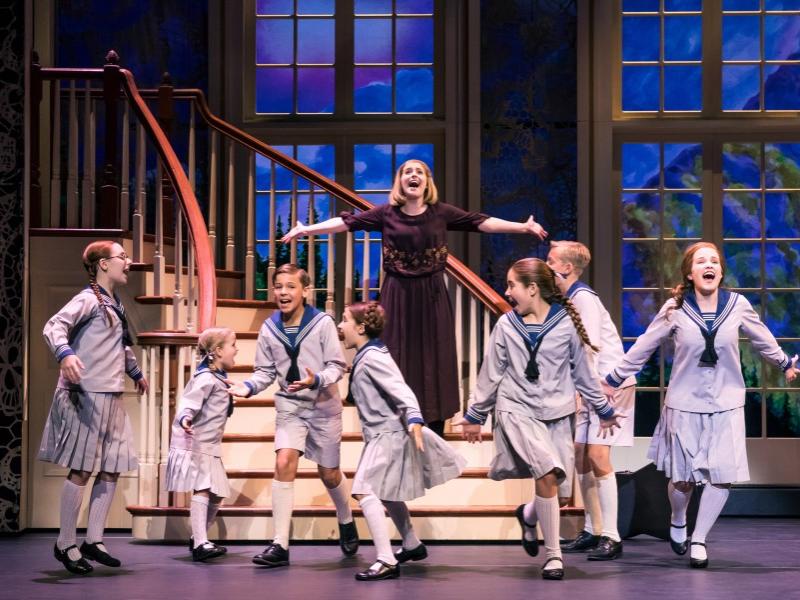 A woman stands at the bottom of a set of stairs with her arms outstretched while a group of young children frolic in front of her.