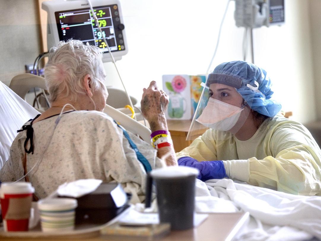 Abigail Adelsberger wears protective gear and kneels by the bedside of Benita Wagner, who is sitting up in a hospital bed facing her.