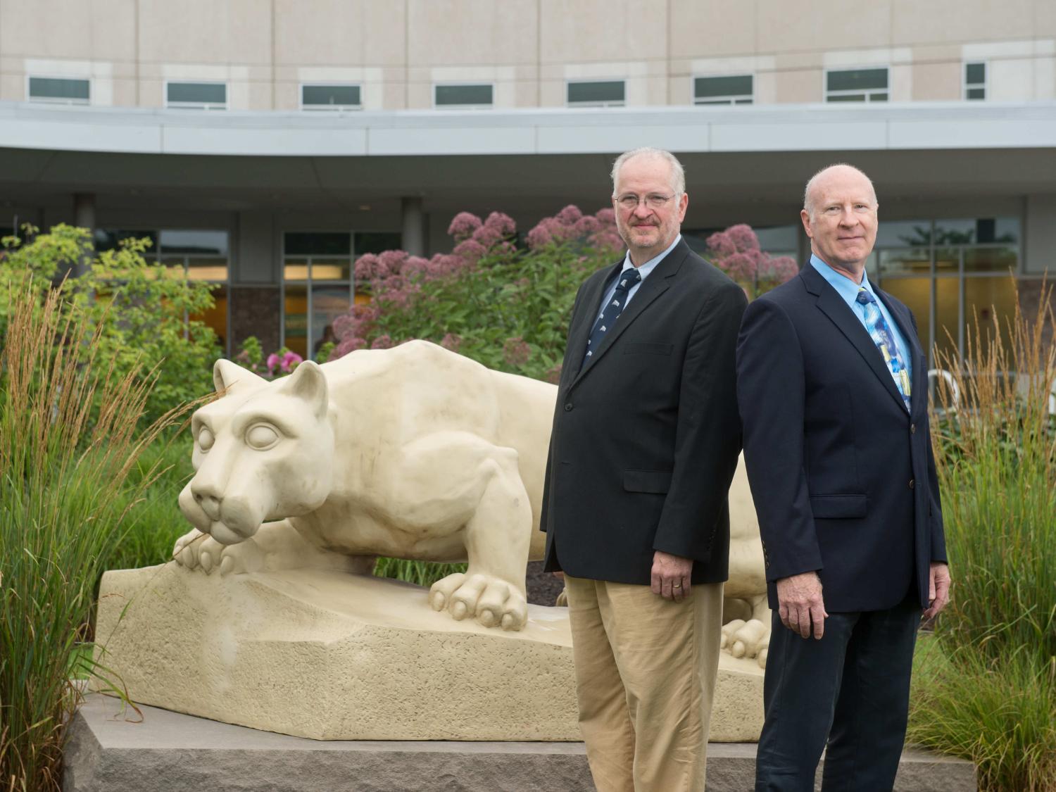 Two men in suits stand back-to-back, slightly off center to the right. A Penn State Nittany Lion statue is at left, and a building is in the background.