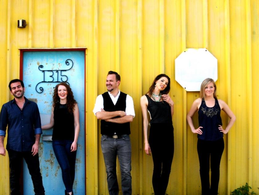 Five people smile while they lean back against a building.
