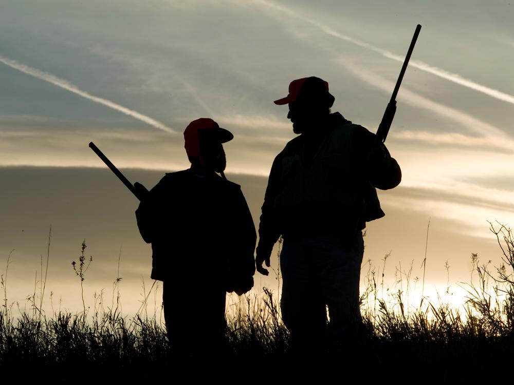 A silhouette of a man and a boy, each holding guns, standing in a field side-by-side. The sky is blue and gray, with some jet airplane vapor trails visible.