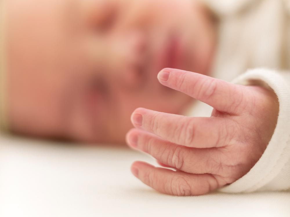 A close-up of an infant’s hand. The baby’s face is seen in the background, out of focus. The baby is sleeping.