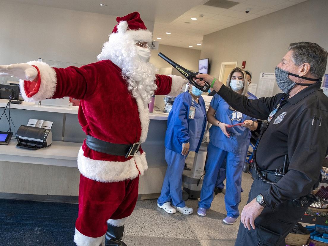 A man in a Santa Claus suit is wanded by a security guard while people in scrubs and surgical masks look on.