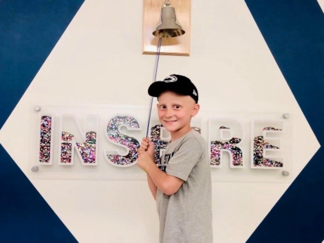 A boy wearing a baseball cap faces the camera and smiles, standing in front of a bell that hangs on a wall along with the word "INSPIRE"