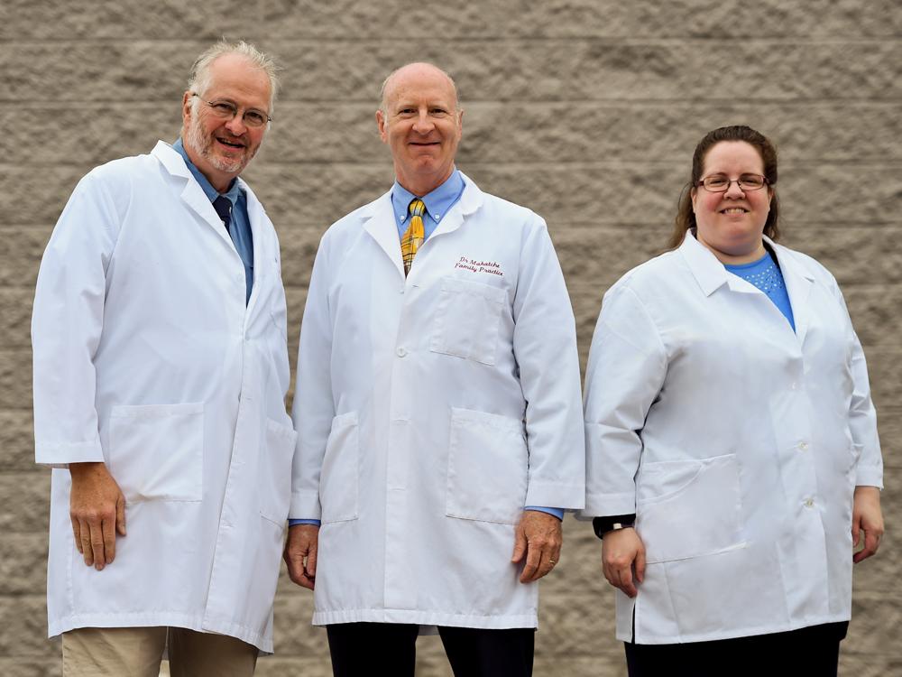 Three people -- two men and one woman -- stand in front of a beige, textured wall, posing for a photo. They're each wearing white clinical jackets.