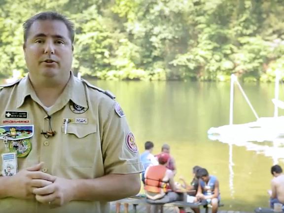 A young man in a beige Boy Scout uniform speaks to the camera. In the background is a lake with about eight people sitting at the water's edge.