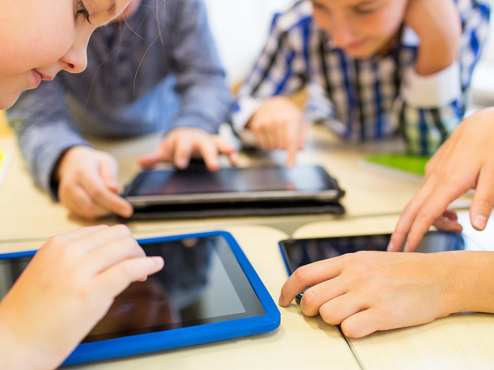 A close-up of four children huddled around a table sharing three tablet computers.