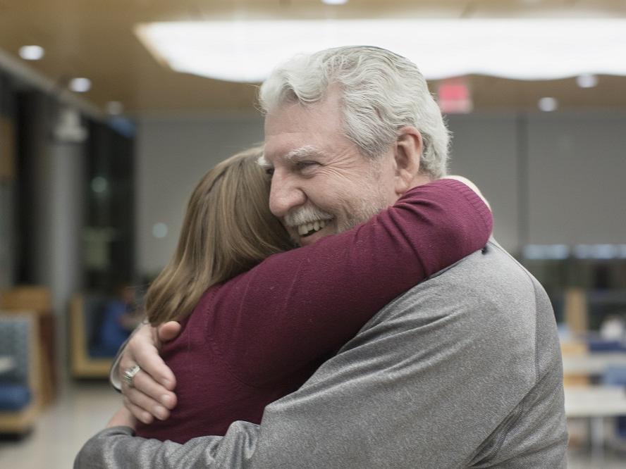 Ed Frederick hugs Dr. Elizabeth Werley at the first “See Me Now” program. Ed has white hair and a moustache and is wearing a gray sweatshirt. Dr. Werley is wearing a long-sleeved sweater. Behind them are cafeteria tables and chairs.