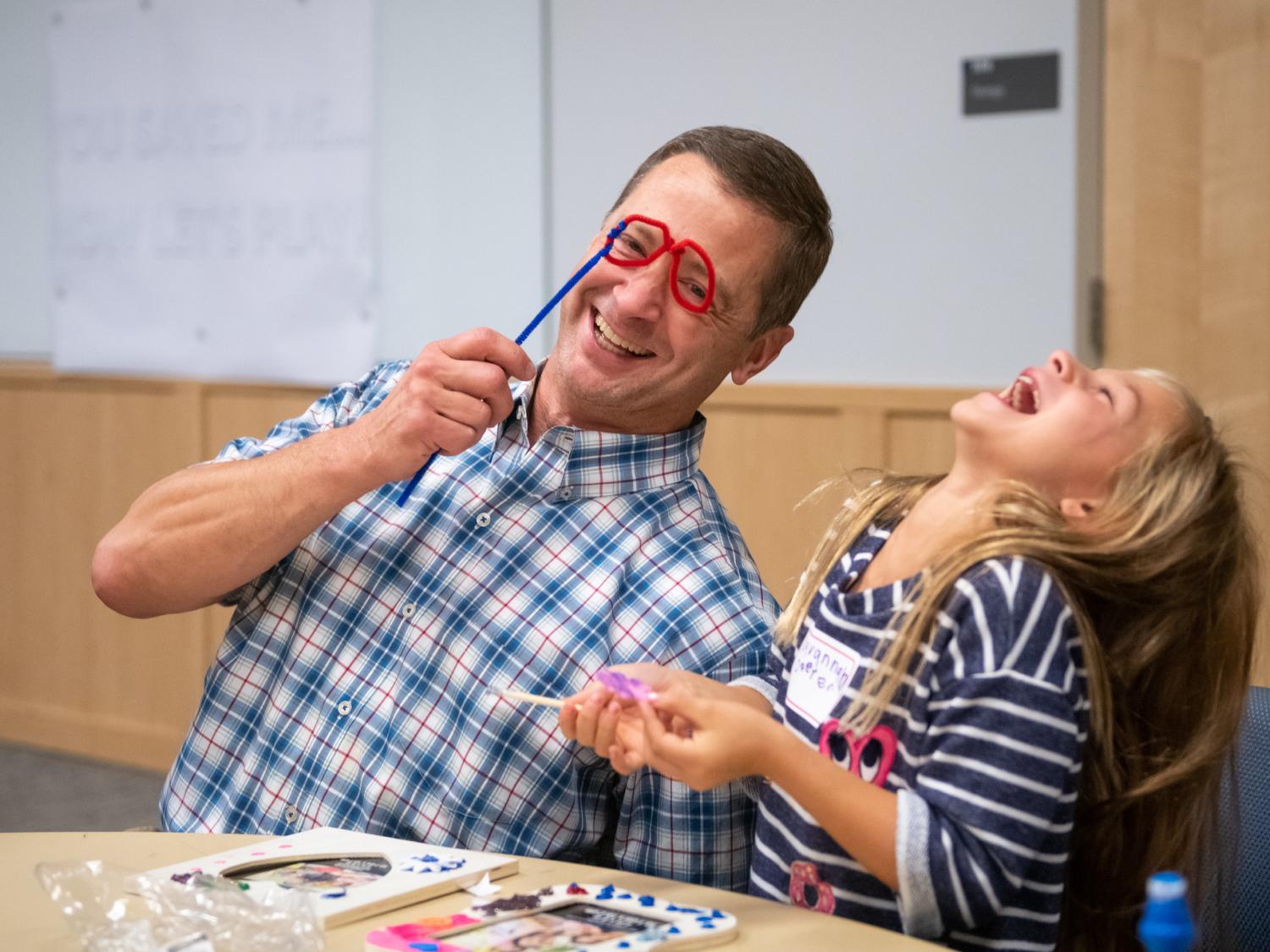 Dr. Christopher O’Hara holds up pipe cleaners twisted into two circles on a straight length of pipe cleaner and leans toward Savannah Doster, who throws back her head and laughs. The two are seated at a table upon which are frames and craft materials.