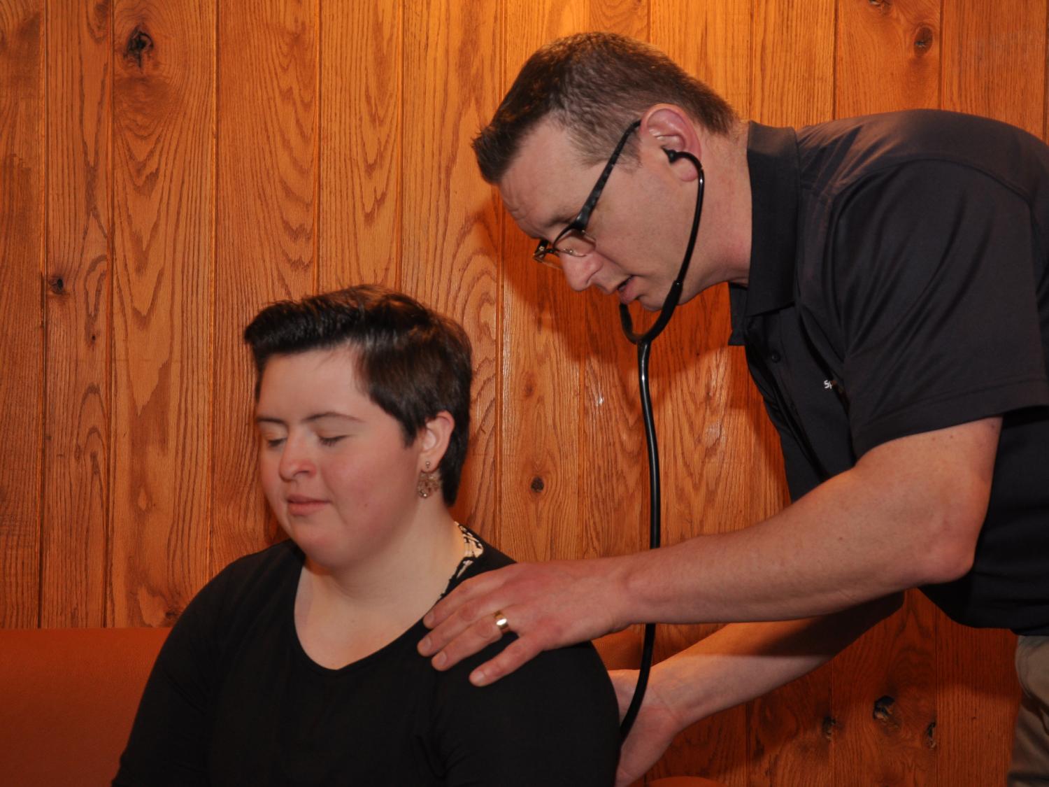 Dr. Peter Seidenberg examines a young woman athlete during the Special Olympics Pennsylvania Winter Games Feb. 10-12. He has a stethoscope in his ears and is holding the diaphragm to her back.