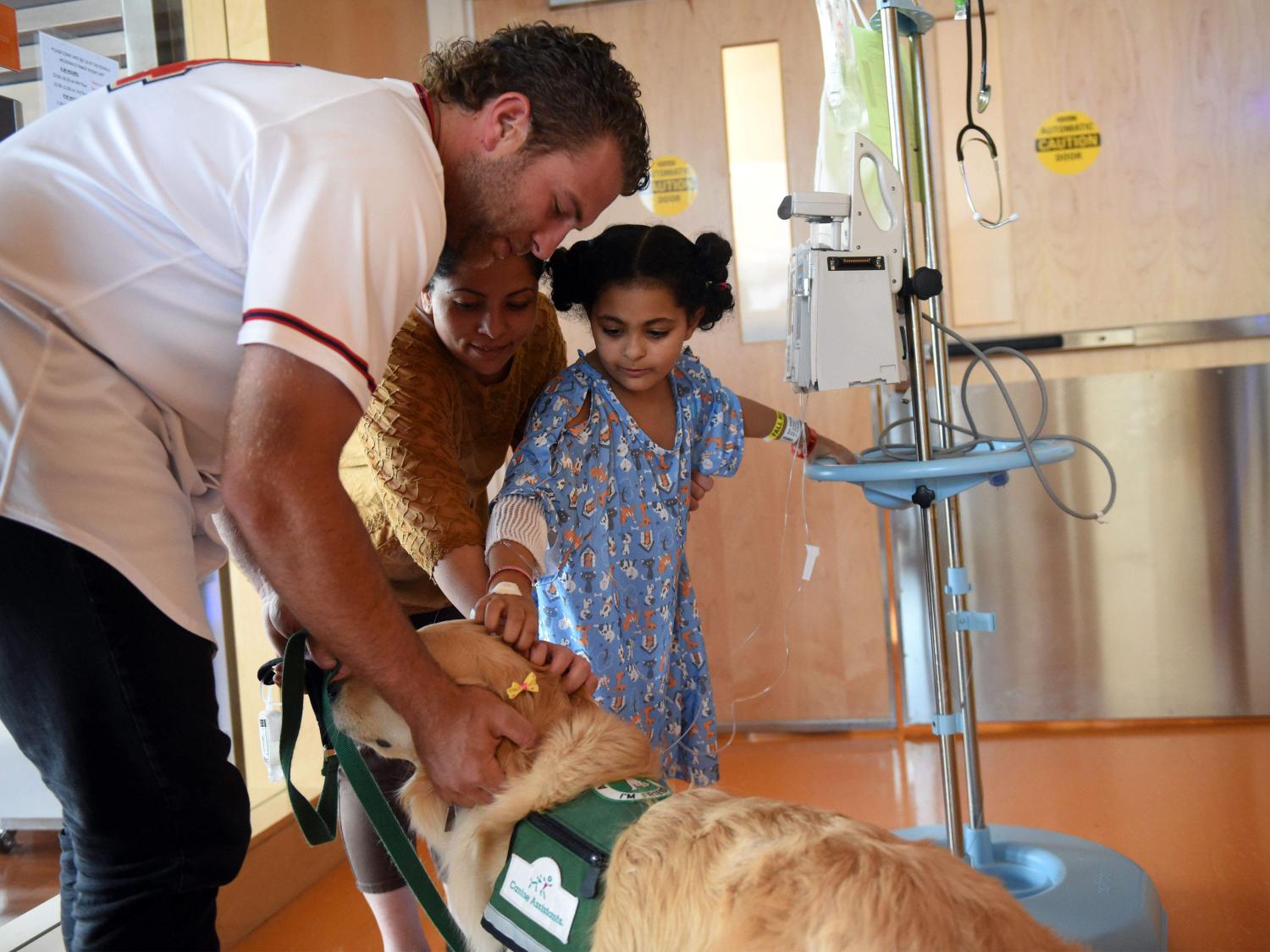 o	A man, woman and young girl pet a service dog in a hospital hallway. A portable cart with IV equipment is next to the girl.