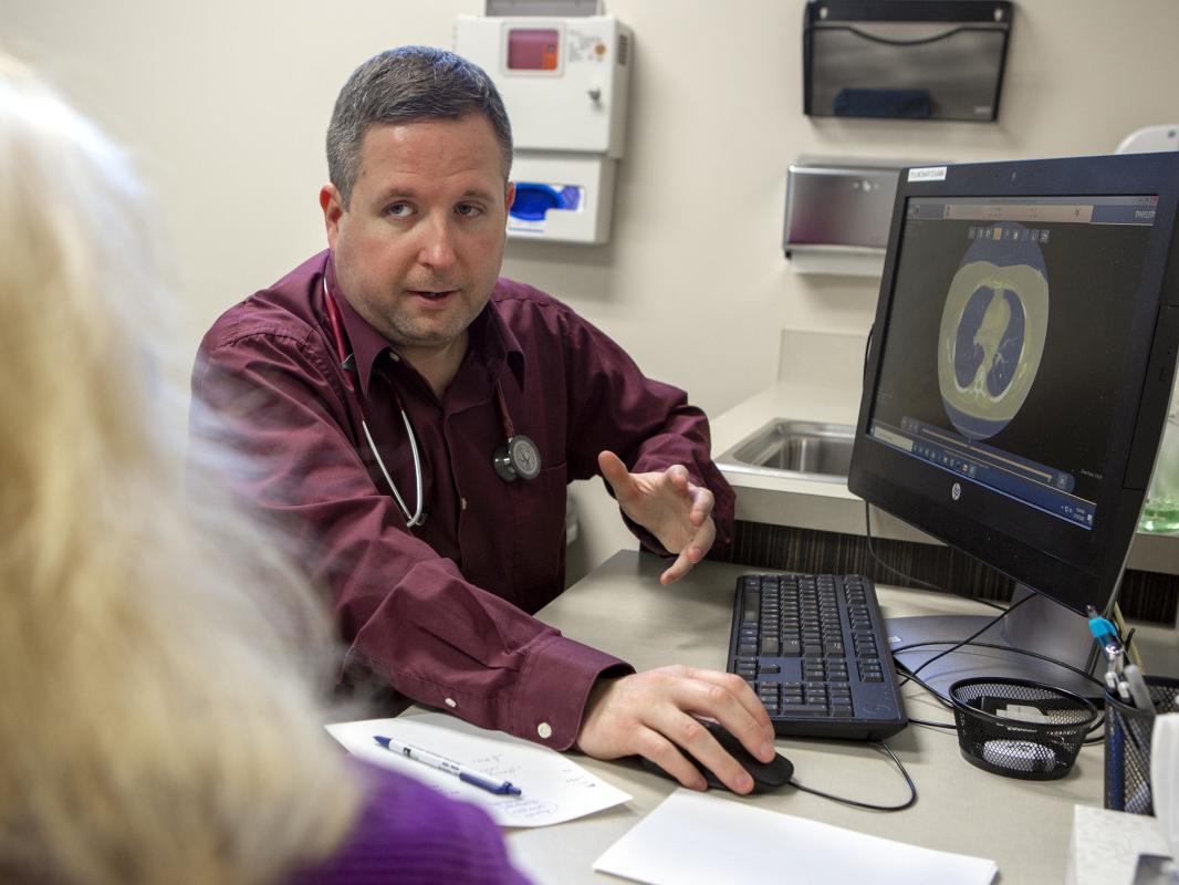 A man in a dress shirt with a stethoscope around his neck sits at a computer in a clinic room. He talks with a woman who is in the foreground, facing away from the camera. A lung scan is on the computer screen.
