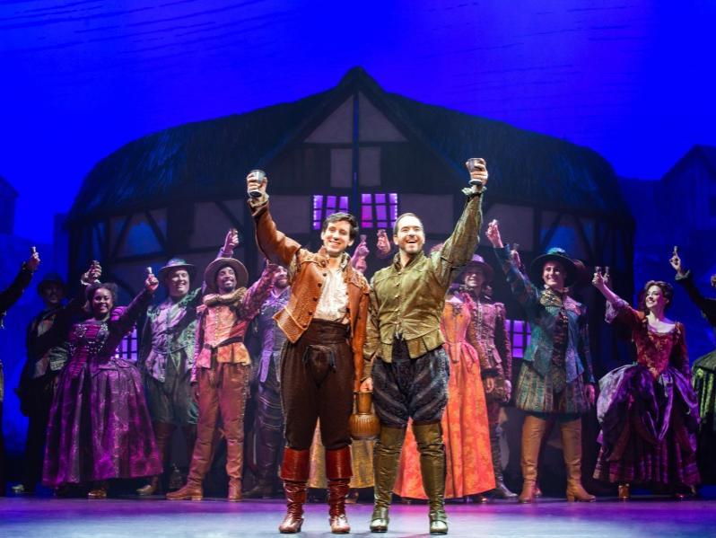 Two men dressed in medieval-style clothing each hoist a chalice in celebration while standing in front of a crowd of costumed actors.