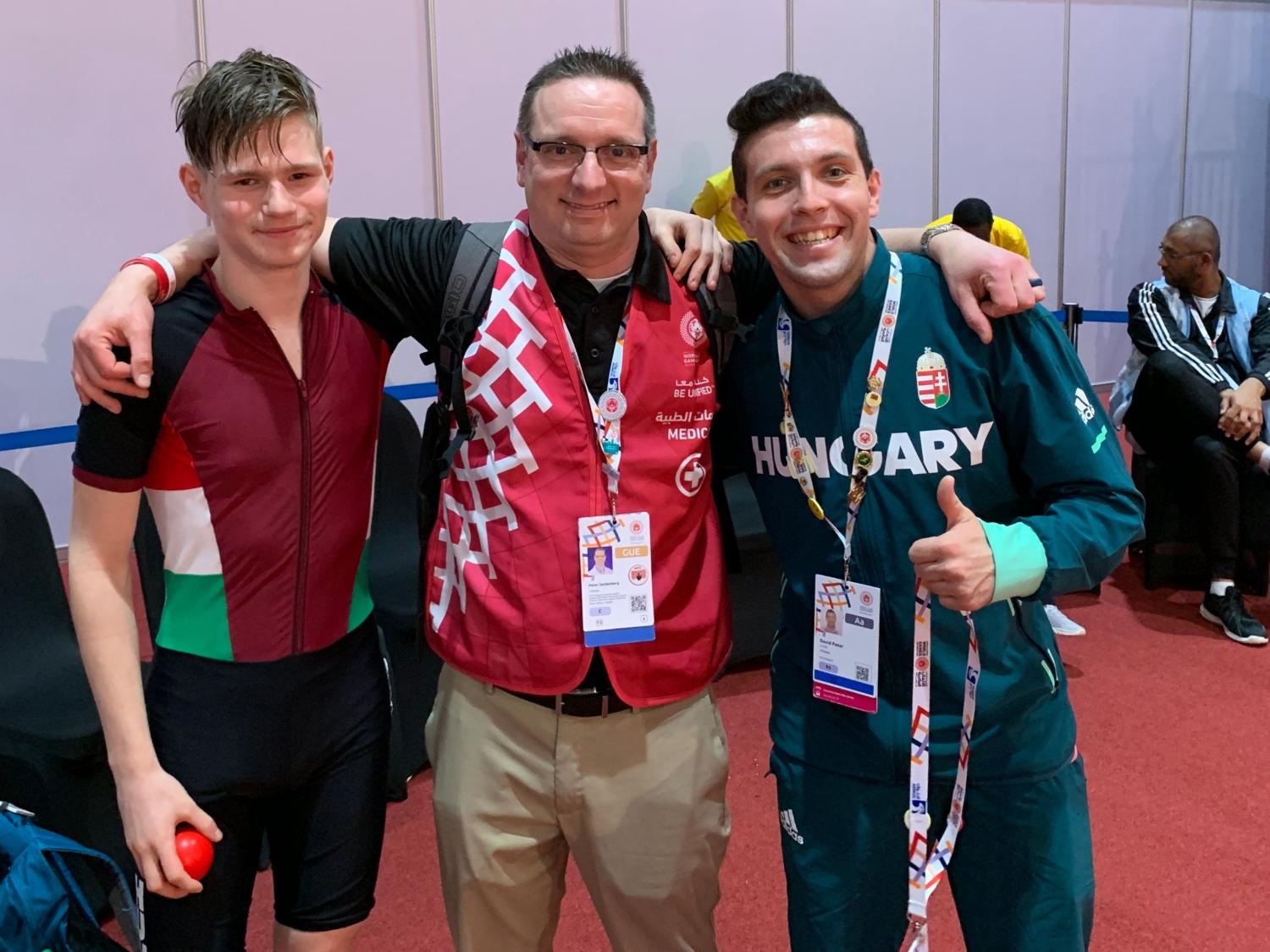 Dr. Peter Seidenberg poses for a photo with two young men who are members of the Hungarian roller skating team. He is wearing a red vest with “Medic” on the right side. The players on either side of him are wearing athletic apparel. 