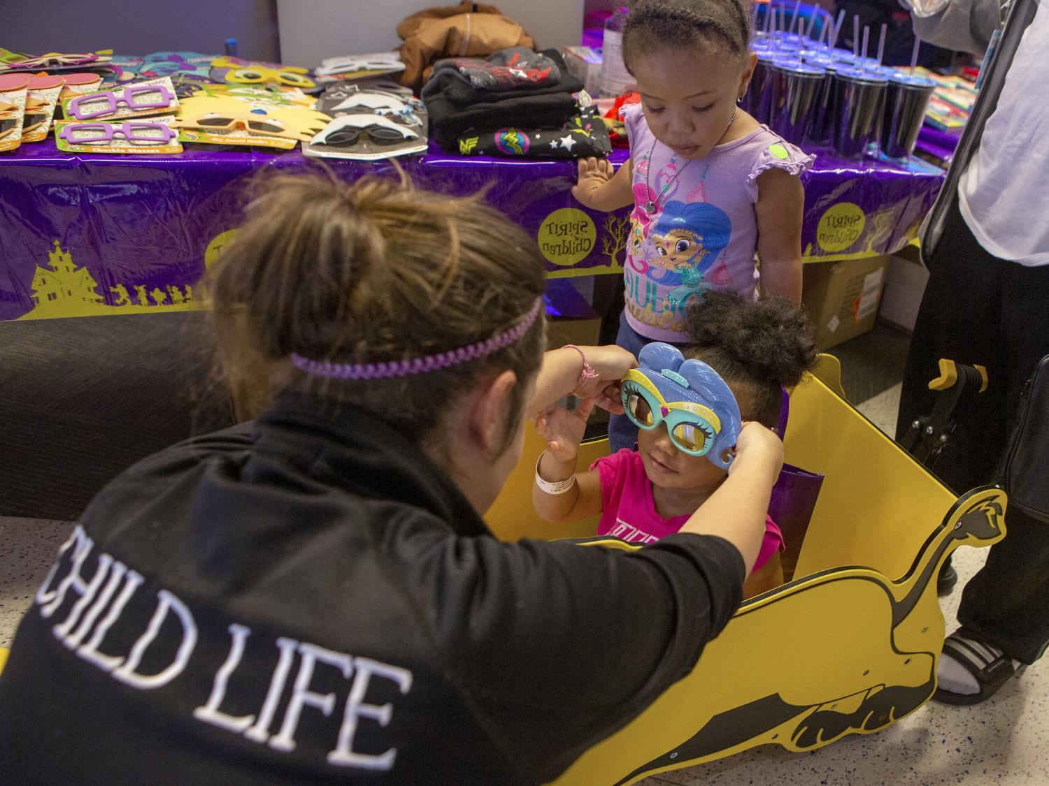 Child Life Assistant Tiffaney Horner places special glasses on the face of 2-year-old Ava Lawson as her sister Aurora, 3, looks on. Ava is seated in a toy car. In the near background is a table with sunglasses, shirts, cups and other merchandise on it.