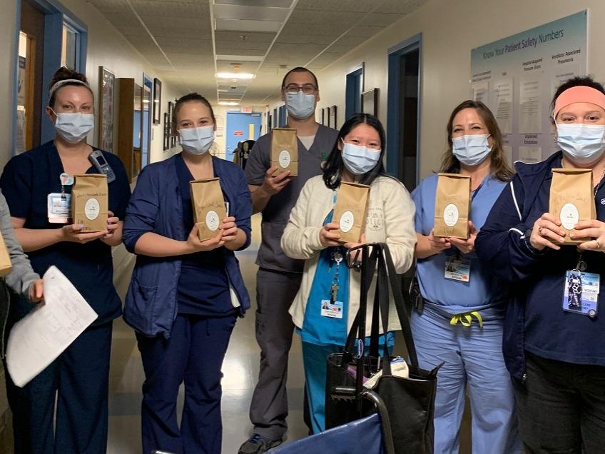 Seven members of the Neonatal Intensive Care Unit are holding bags of coffee. They are all wearing scrubs and masks and standing in the hallway of their unit.