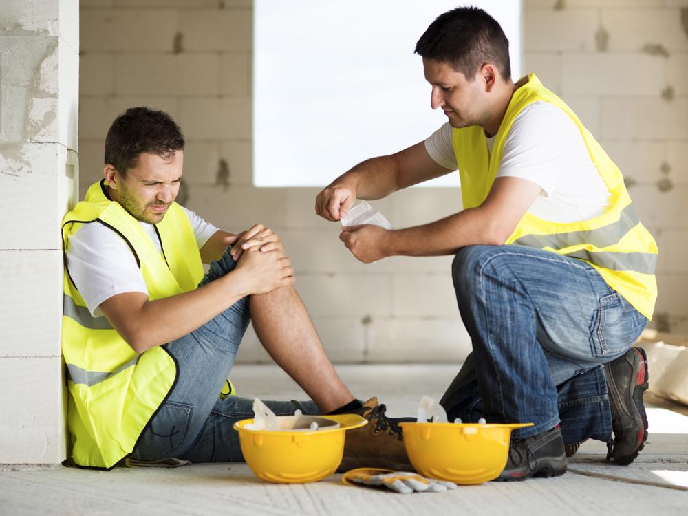 A man in a yellow construction vest sits on the ground against a wall, holding his knee in apparent pain. Another main in a construction vest kneels next to him, preparing to apply a bandage.