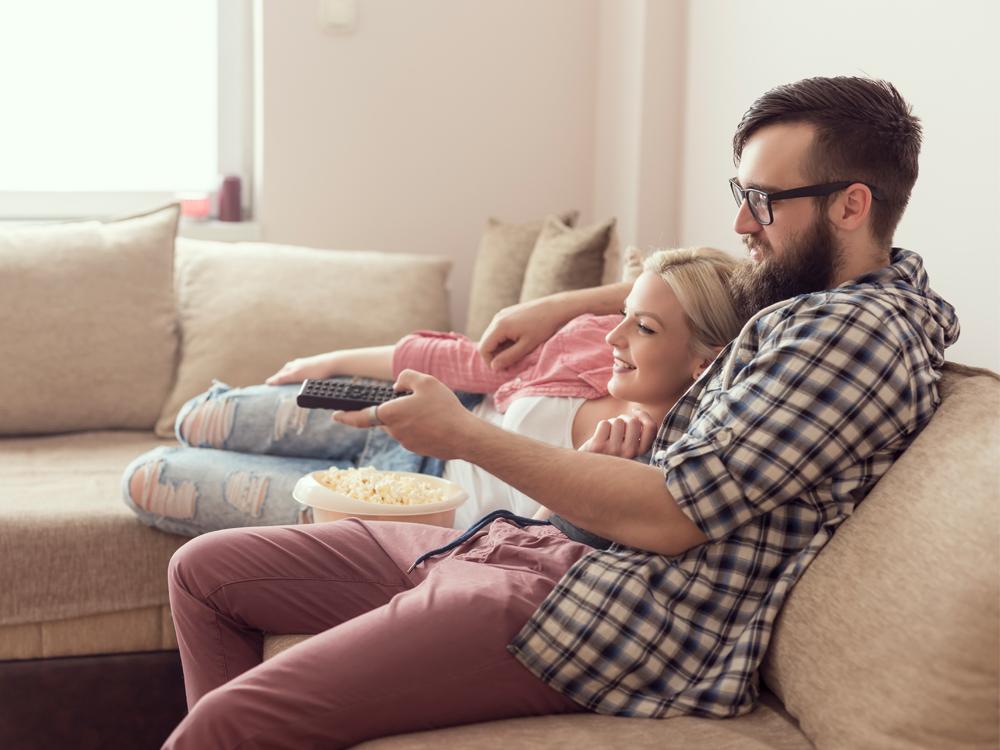 Side view of a young man and young woman, both in their 20s, sitting on a couch. The man has his arm around the woman and holds a TV remote control in his hand. A bowl of popcorn is in his lap.
