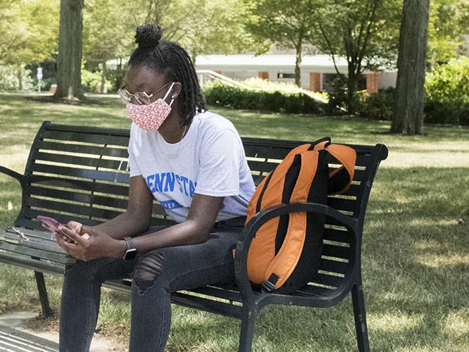 A Penn State student wearing a face mask looks at her phone while sitting on a campus bench.