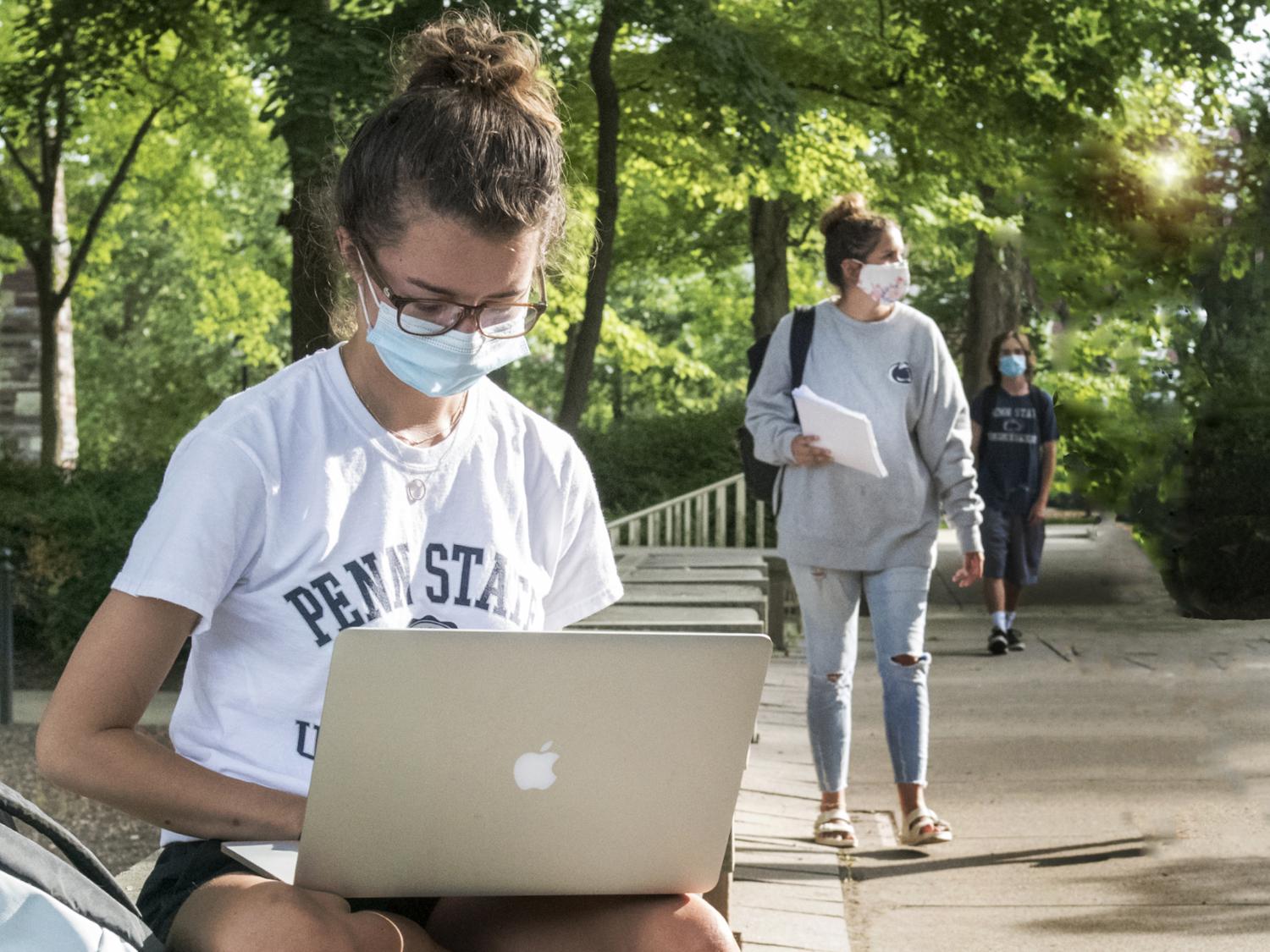 A student wearing a mask sits with a laptop outside of Old Main on Penn State's University Park campus. 