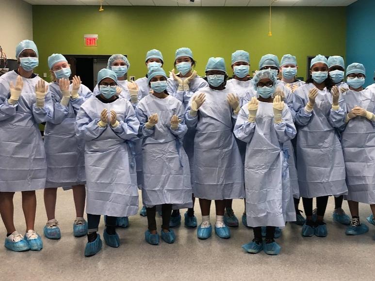 Nineteen high school students stand together in a room with a green wall wearing blue surgical gowns, masks and hair and shoe coverings. They are wearing surgical gloves and holding up their hands.