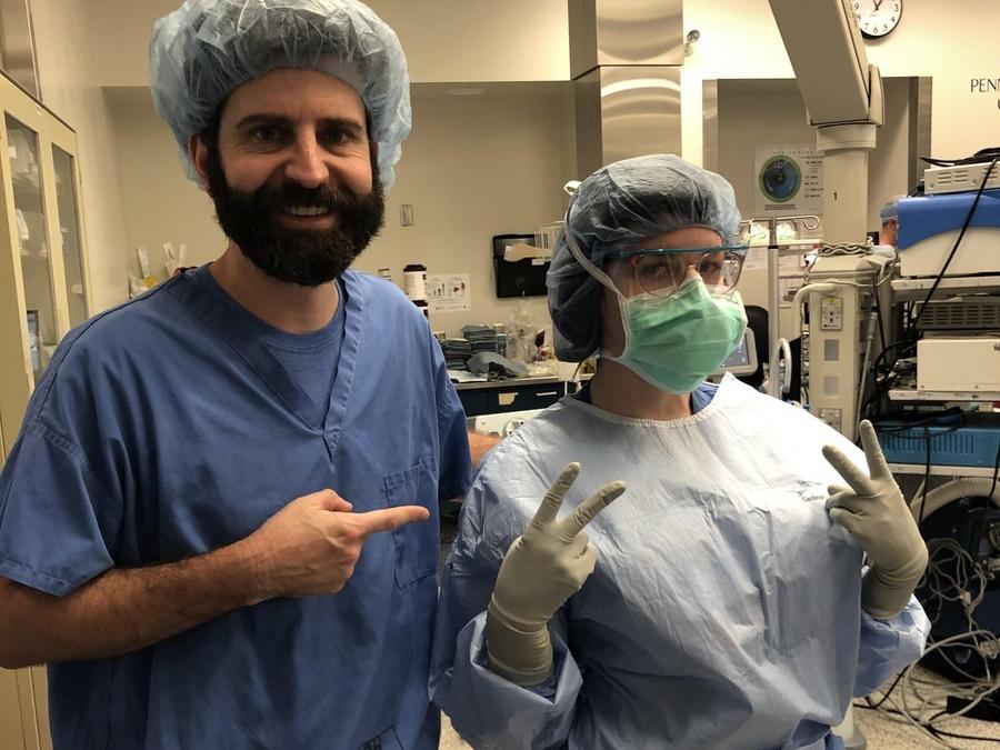 Dr. Eric Pauli poses with registered nurse Kiersten Marie in the operating room. Marie wears a surgical gown, gloves, mask, hair net and glasses. Pauli, who has a beard and moustache, wears scrubs and points his index finger toward Marie.