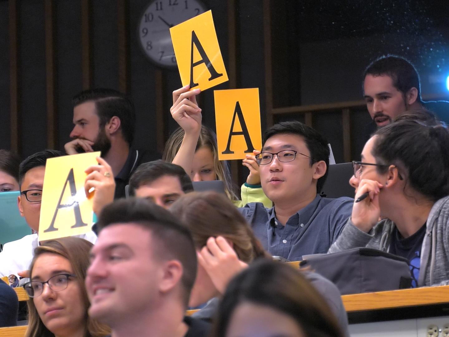 Several students are seated in a tiered classroom. Some of them hold up cards with the letter “A” on them.
