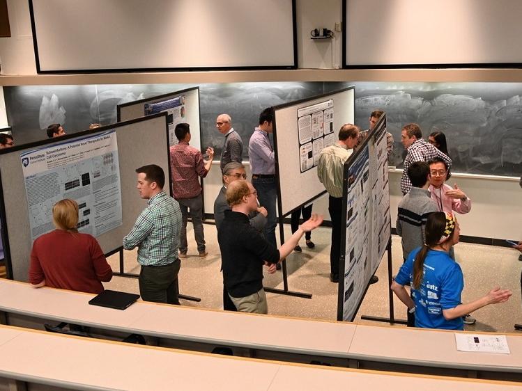 Men and women stand at the front of a classroom and talk around easels displaying posters. Behind them, a blackboard stretches across the room.