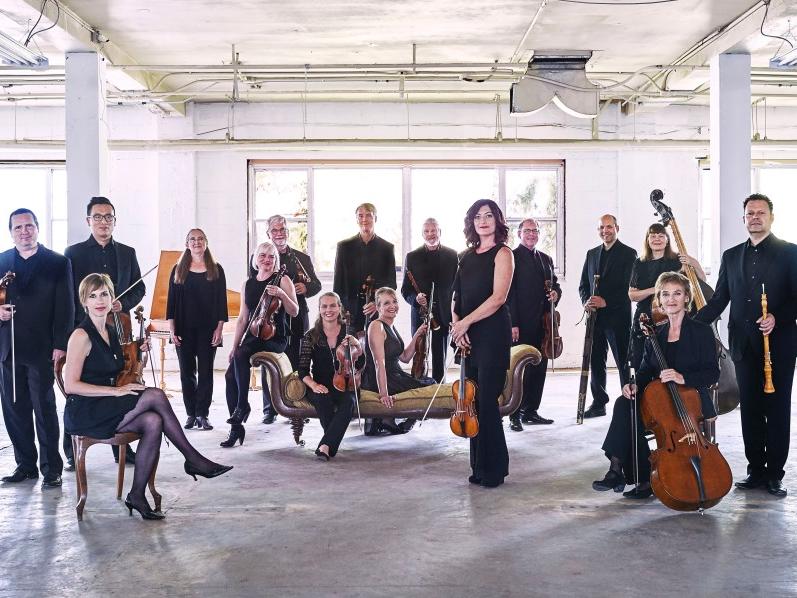 A group of musicians holding their instruments stand in a cluster in a well-lit warehouse space.