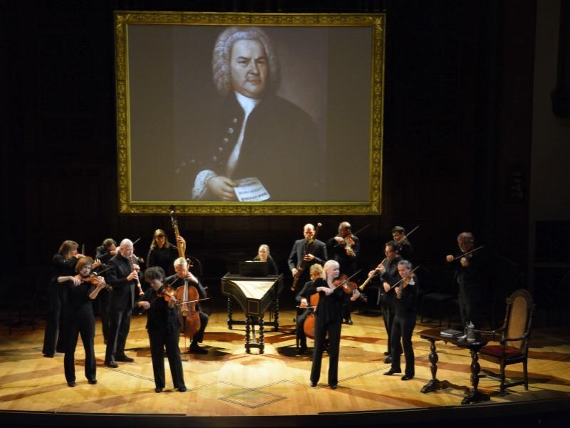 Tafelmusik Baroque Orchestra musicians play their instruments on stage in front of a backdrop featuring an image of J. S. Bach.