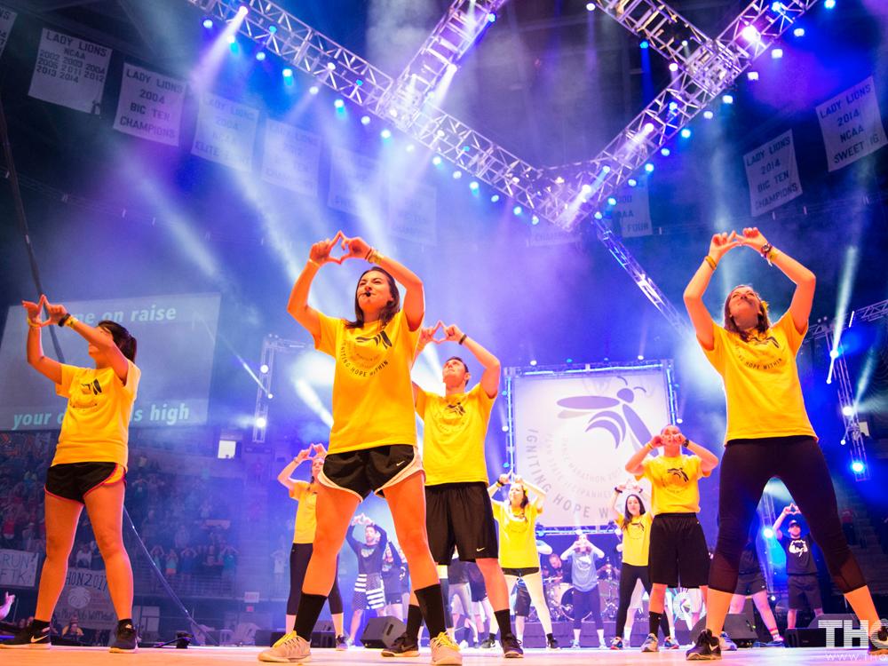 Several college students in yellow shirts and black pants and shorts stand on a stage, holding their hands high and making a diamond symbol with four fingers. Purple lights shine from above.