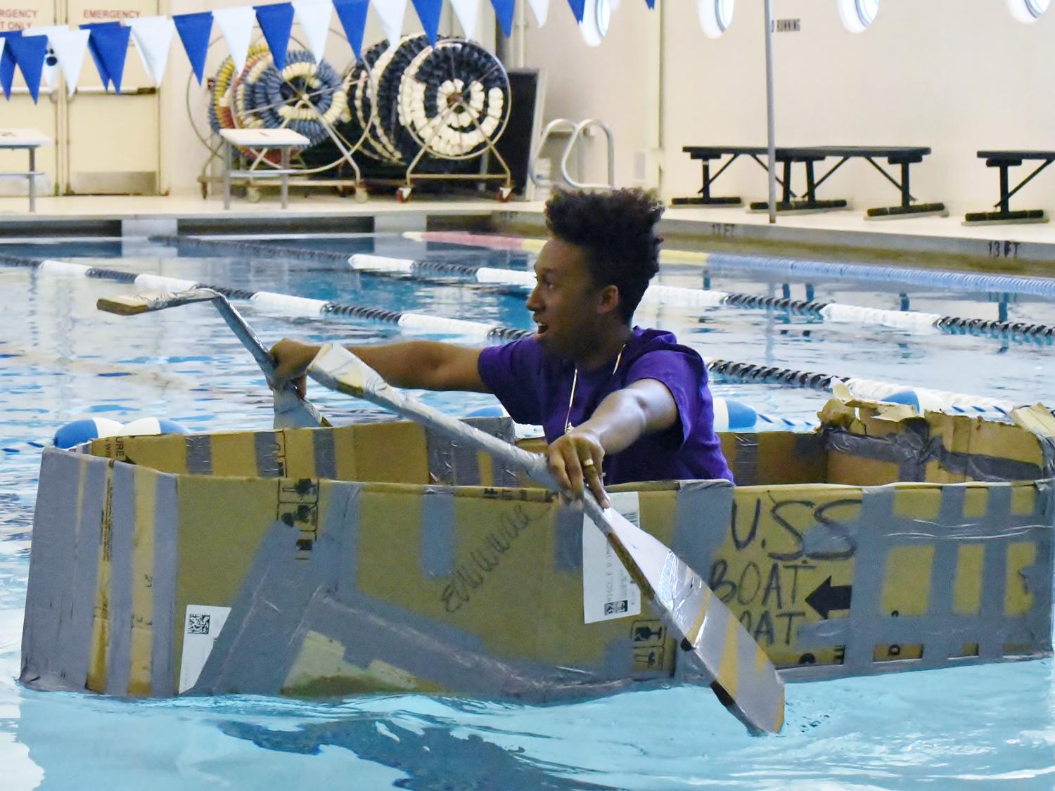 A high school student sails a cardboard boat in a swimming pool.