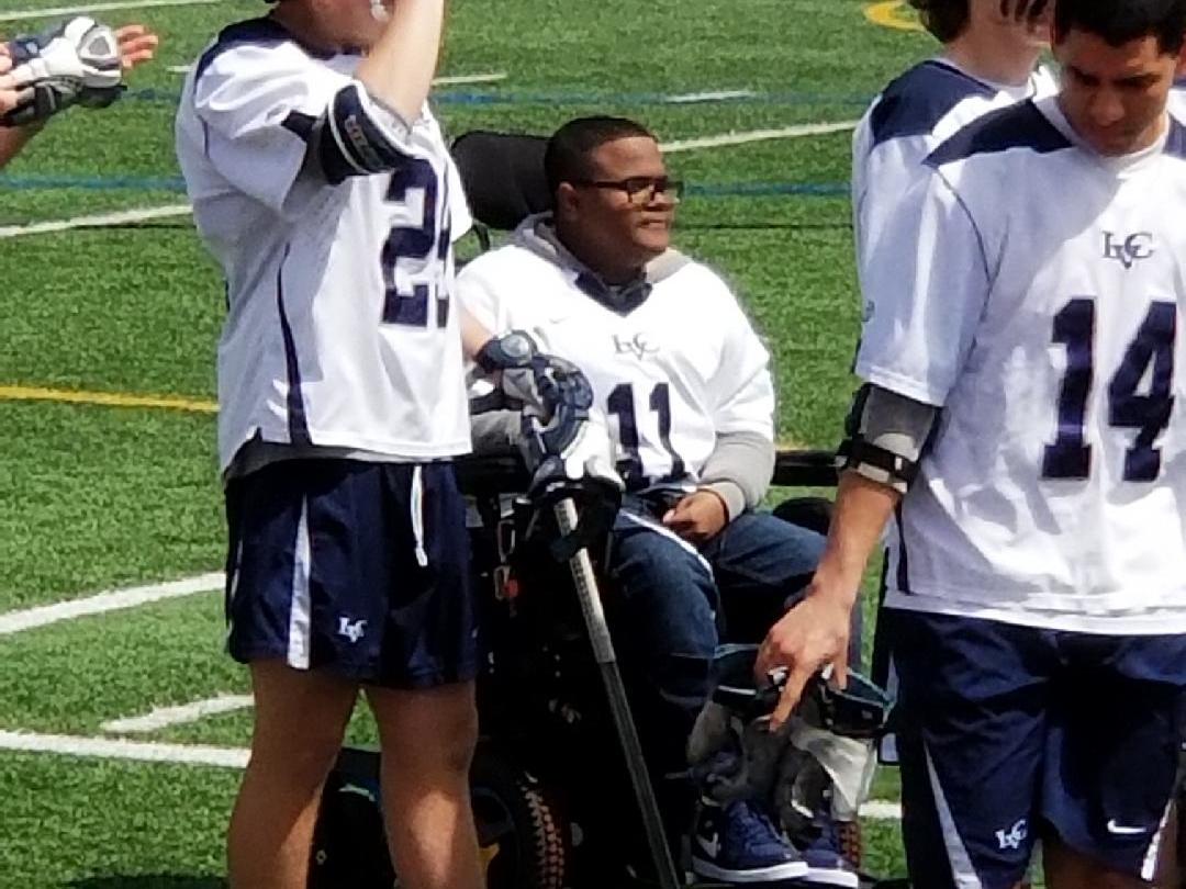 A young man in a wheelchair is on a playing field, with a few lacrosse players walking along side.