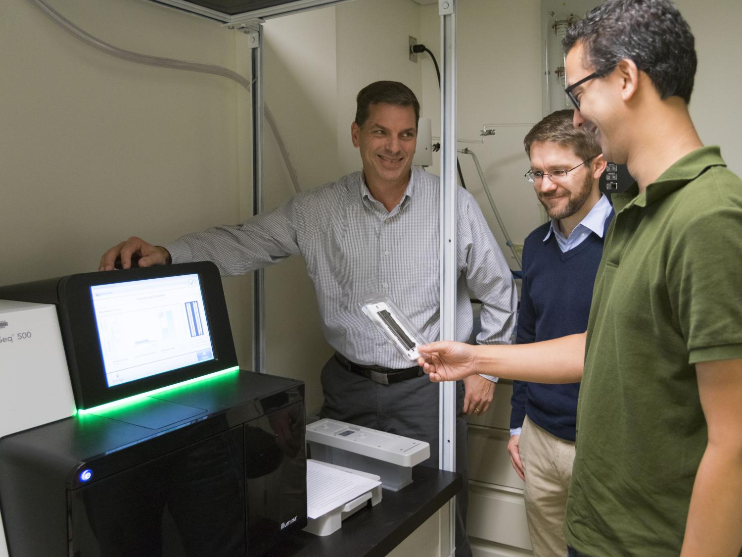 Three researchers gathered around a gene sequencer smiling