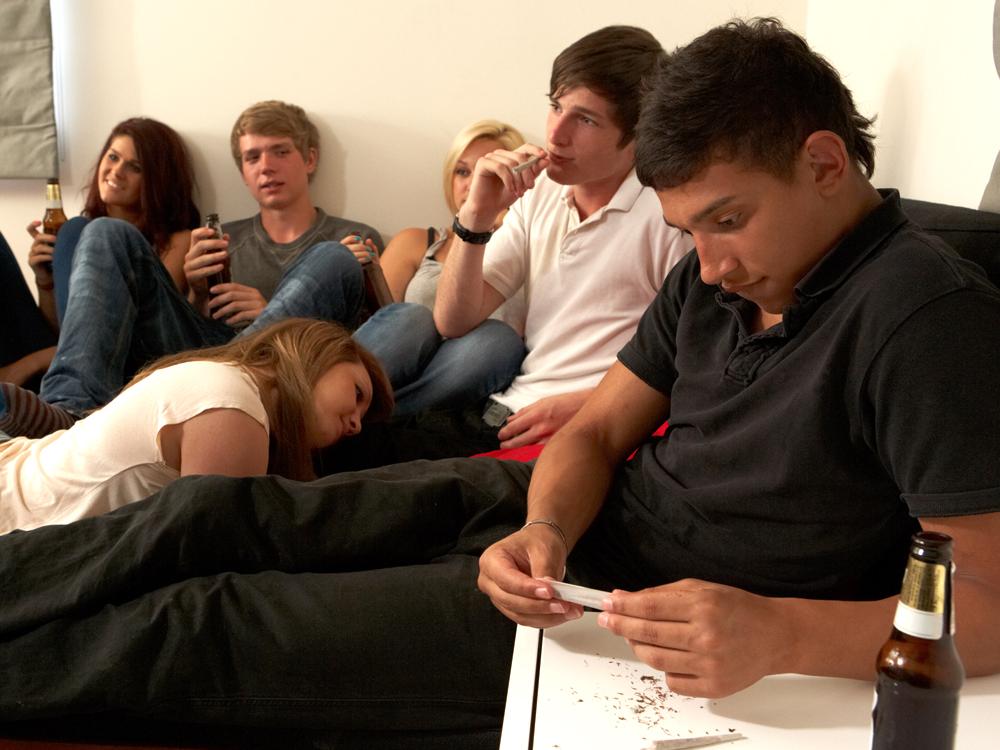 Six teenagers sit around a room. The teen closest to the camera is rolling a joint, another is smoking a cigarette, and two others are holding beer bottles.