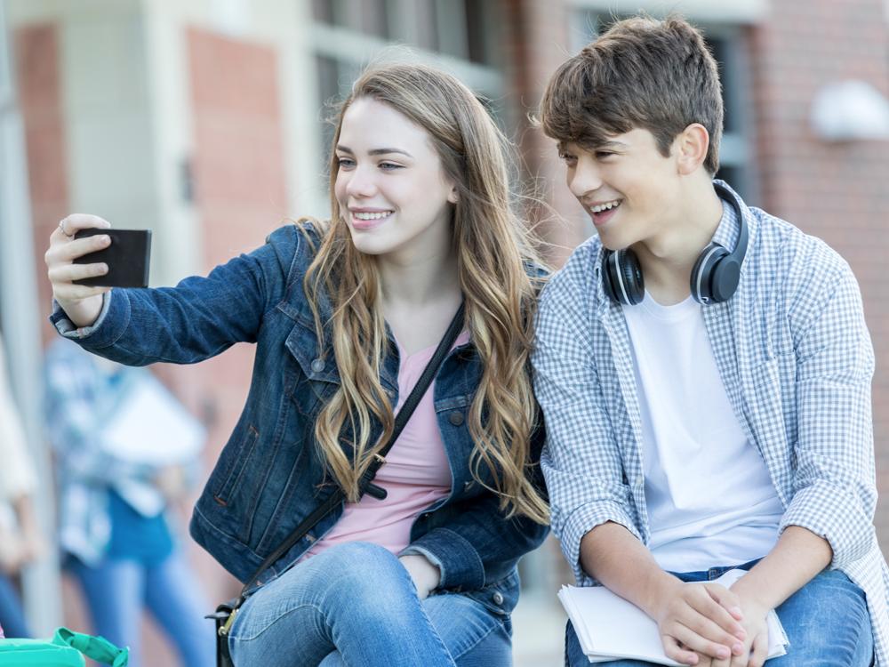 A teenage boy and girl sit together and look into a phone the girl is holding up, apparently posing for a selfie. A brick building with windows is in the background.