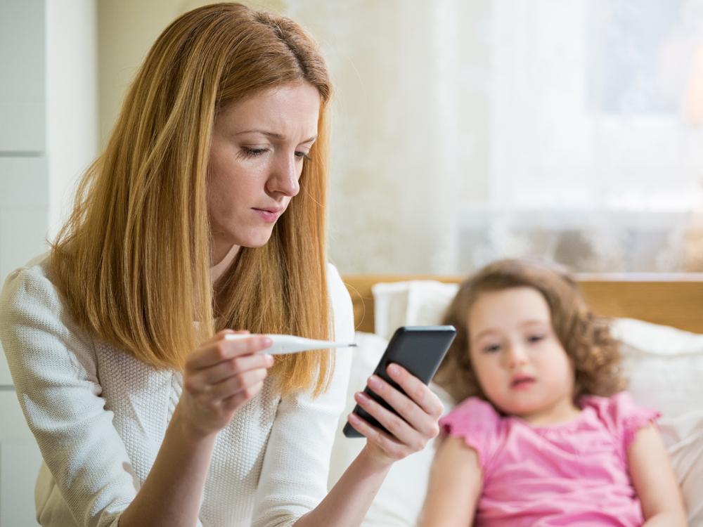 In the foreground, a woman holds a digital thermometer and looks down at her phone, in the other hand. In the background, a young girl in a pink shirt lies in bed.