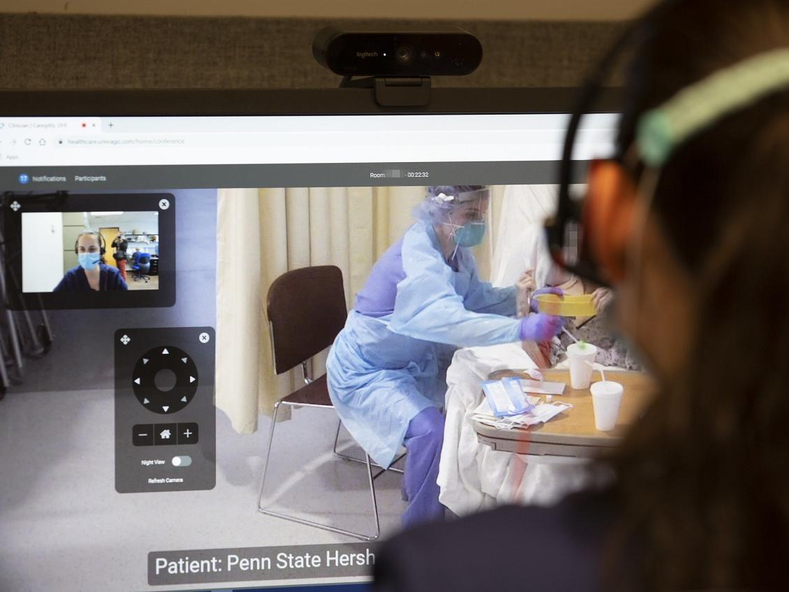 Image shows over the shoulder someone looking at a computer monitor. In one window on the monitor is the view of someone in a mask. To the right, someone in personal protective equipment works with a patient.