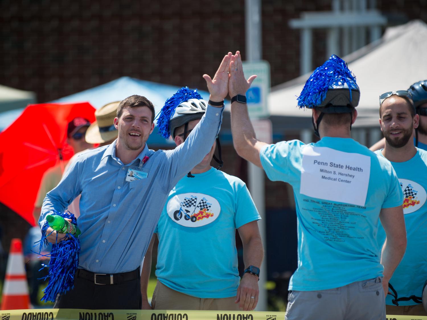 Joe Connors in a button-up shirt holding a Mountain Dew bottle and a pompom in his right hand, high fives a man in a T shirt and a helmet with a pompom attached to the top.