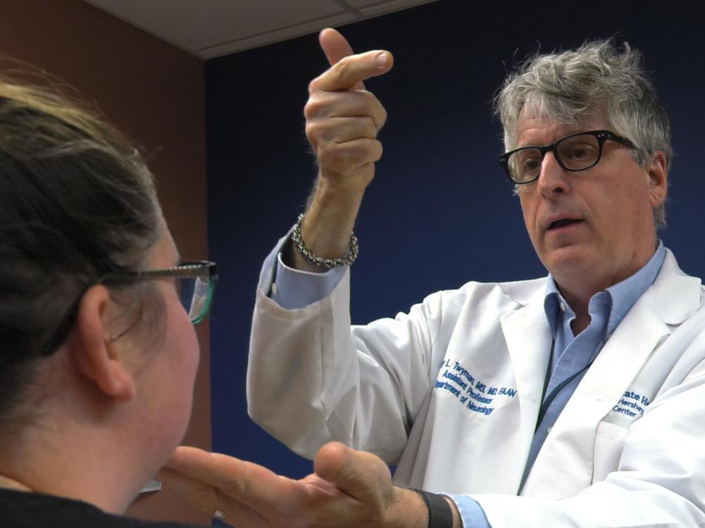 Dr. Cary Twyman, a neurologist at Penn State Health, stands in front of a woman, his left hand on her chin, his right hand extended slightly into the air.