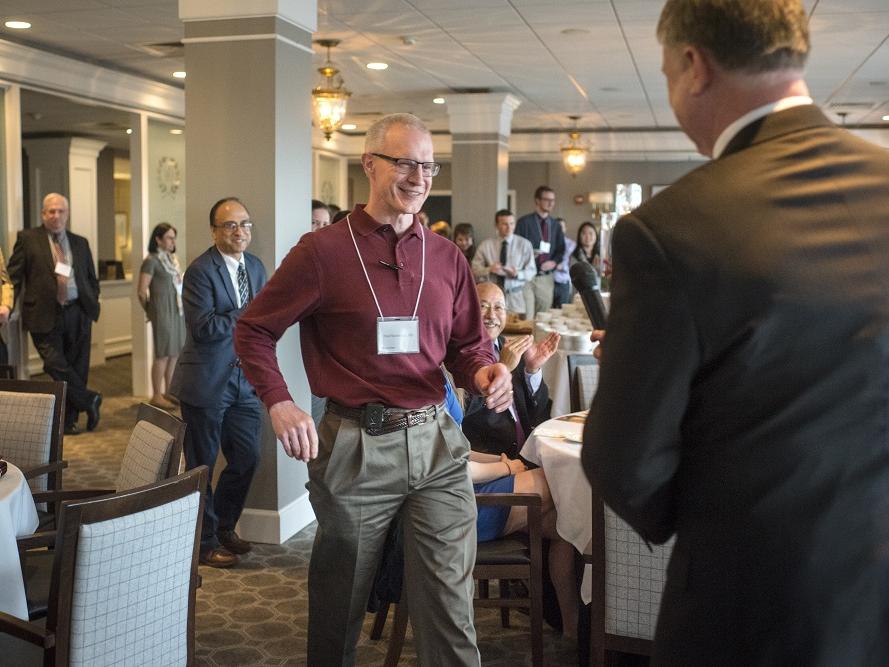 Paul Sherbundy smiles as he walks toward a man in a suit. All around Sherbundy, people in formal business attire smile and applaud or look on. They are in a conference room with upholstered chairs at circular tables.