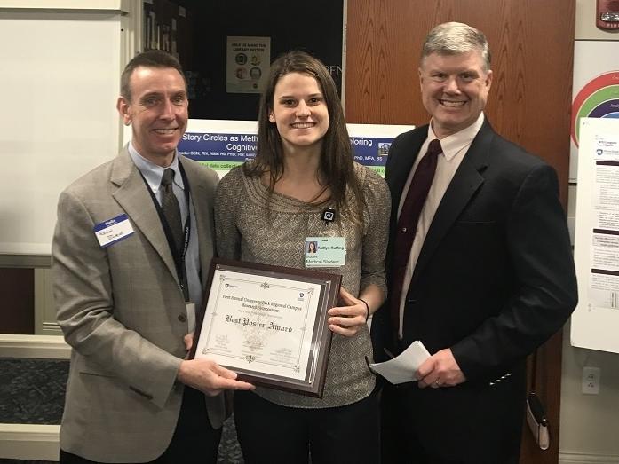 Two men in suits and ties stand on either side of a woman, who holds a plaque, which reads “Best Poster Award.” Three stand in a room surrounded by posters on easels.