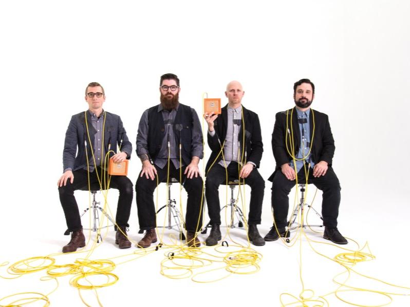 Four men wrapped in electronic cords sit in chairs and look straight ahead.